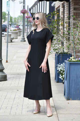 Woman wearing a black dress standing on a sidewalk with plants and street lamps in the background.